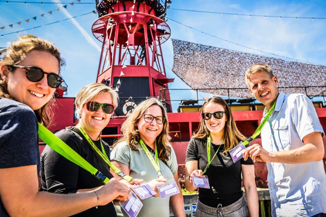 Gruppe von fünf Personen mit Teamkit-Umhängebändern posiert fröhlich vor einem roten Leuchtturm bei sonnigem Wetter.
