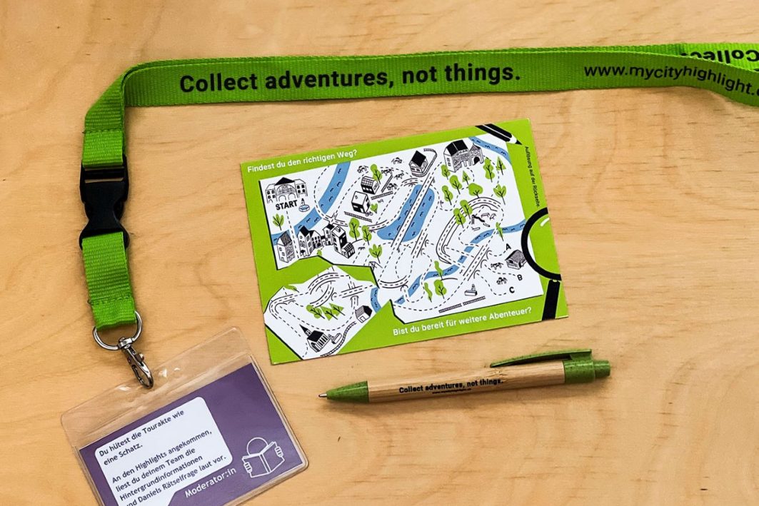 Nahaufname des Teamkits bestehend aus Umhängebändel mit Rollenkarte, vorfrankierter Postkarte sowie Kugelschreiber auf Holztisch liegend.